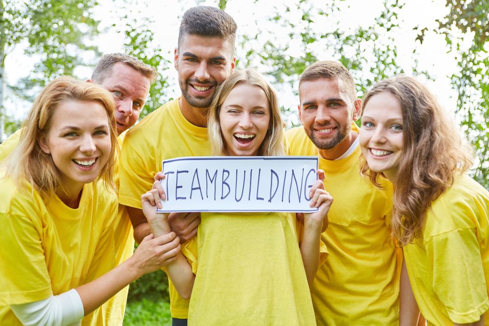 Grupa ljudi u žutim majicama drži znak sa natpisom “Teambuilding” i pozira zajedno u prirodi.